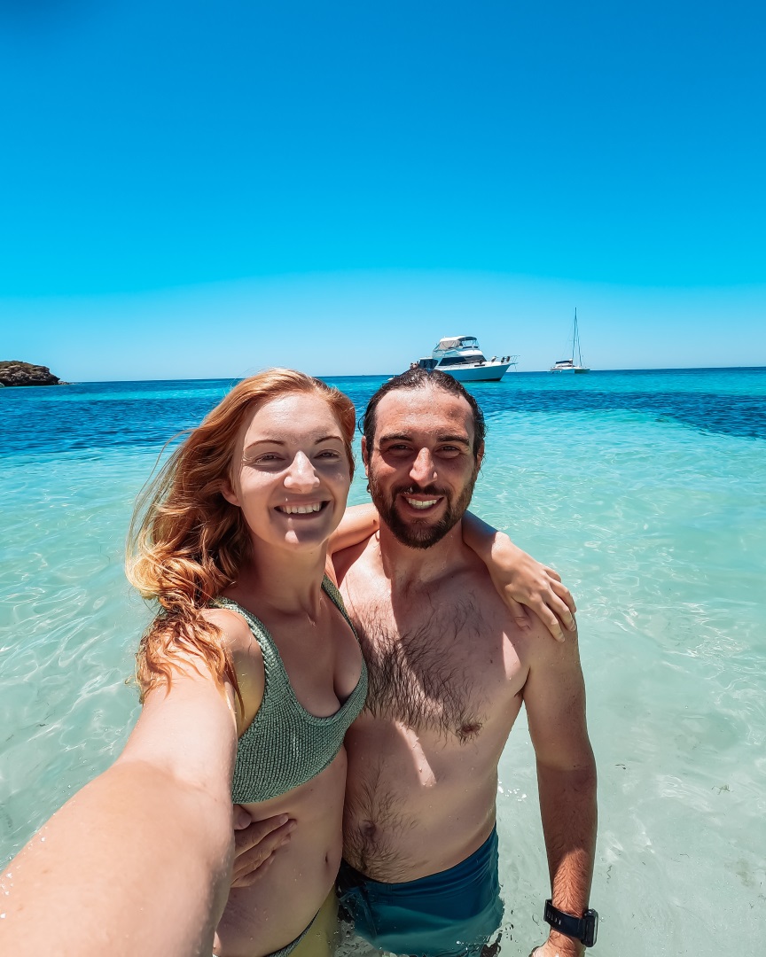 A smiling couple takes a selfie while standing in the shallow, clear waters of Little Parakeet Bay, Rottnest Island. Behind them are ferries to take for the best Rottnest Island tours