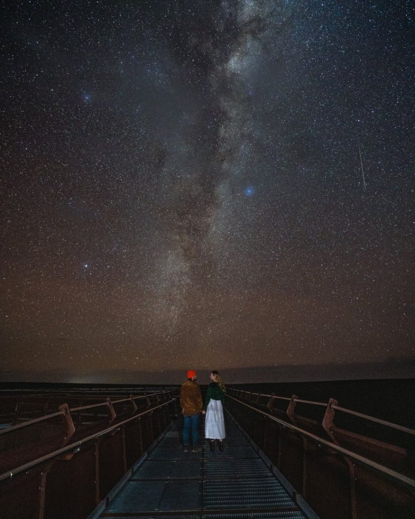 Two people stand on a walkway under a star-filled sky with the Milky Way overhead at the Skywalk in Kalbarri, WA.