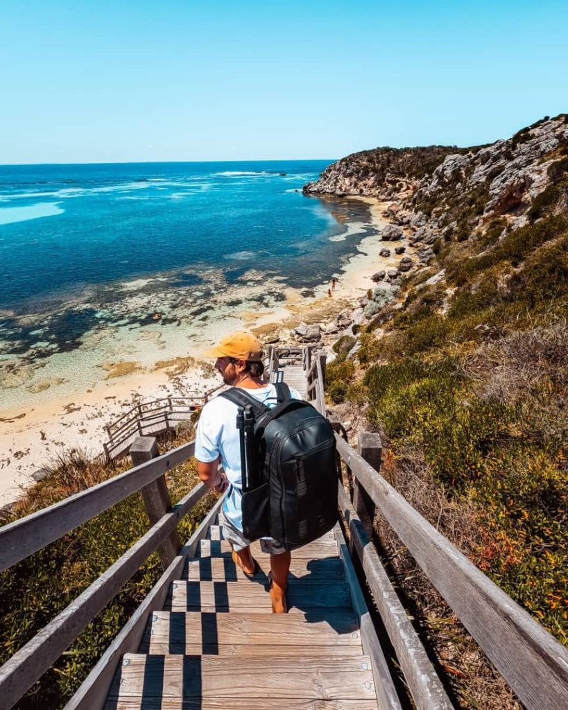A man with a large backpack and a tripod walks down wooden stairs toward a picturesque beach at Parker Point on Rottnest Island Western Australia.