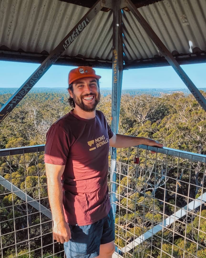 A man wearing a red cap and maroon shirt stands smiling at the top of the Dave Evans Bicentennial Tree lookout in Pemberton, Western Australia. This image highlights things to do Pemberton WA.