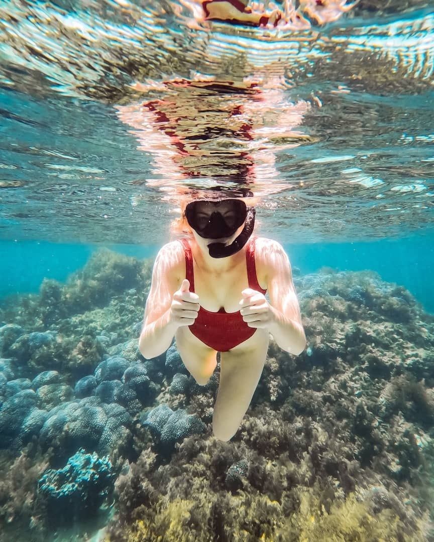 A snorkeller in a red swimsuit and mask gives a thumbs-up while exploring the vibrant coral reefs of Pocillopora Reef, showing what to do on Rottnest Island.
