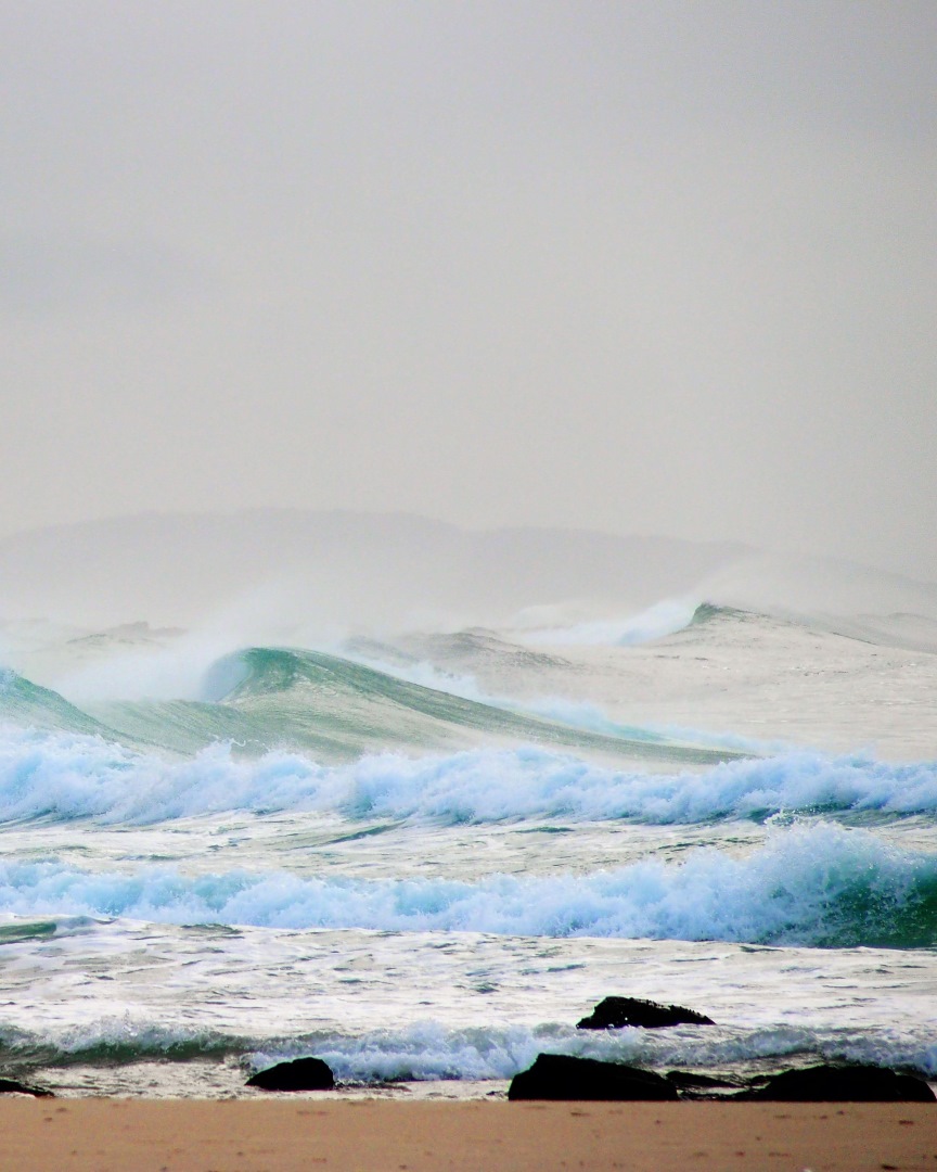 Waves crash against rocks on a sandy shore under an overcast sky. The sea appears rough and choppy, with mist rising in the background.