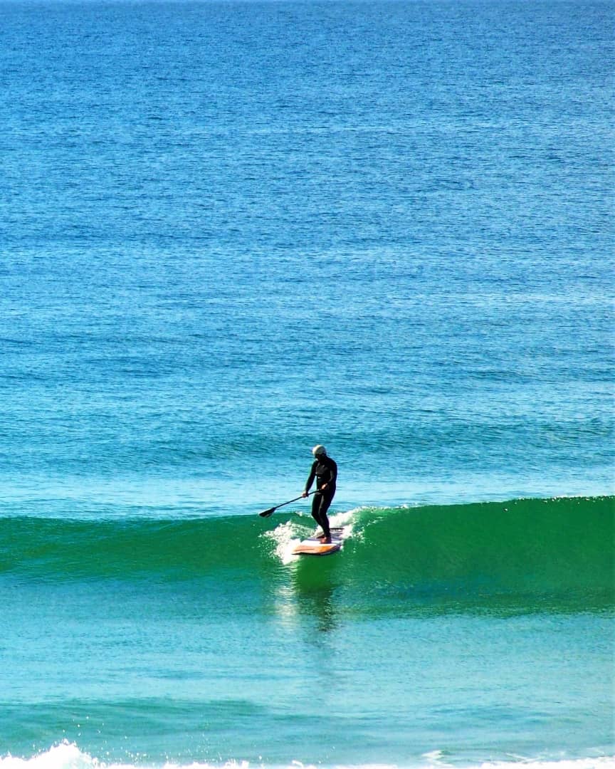 A person is paddleboarding on a clear, blue ocean wave. The individual is wearing a wetsuit and a hat.