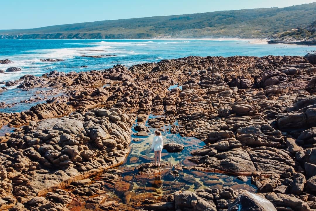 A woman stands on the rocky coastline of Yallingup in Western Australia, surrounded by tide pools with the ocean waves crashing in the background.