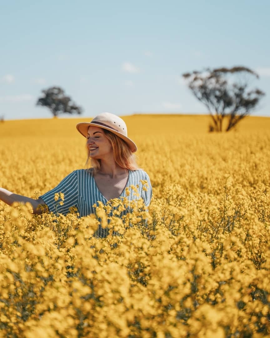 A woman in a striped dress and hat smiles while standing in a vibrant field of yellow wildflowers near Perth, Australia. This scene showcases the best time to visit Perth.