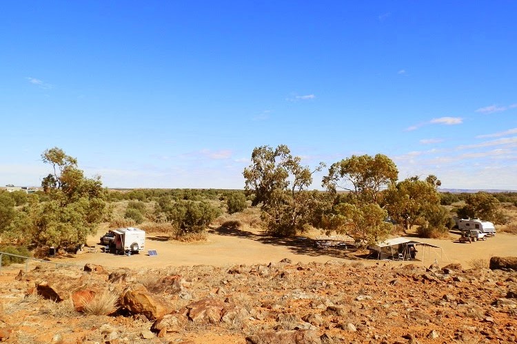 A view of the Farina Campground in Outback South Australia, featuring a few camper trailers and tents set up under sparse trees. The scene shows a dry, rocky landscape with a clear blue sky overhead.