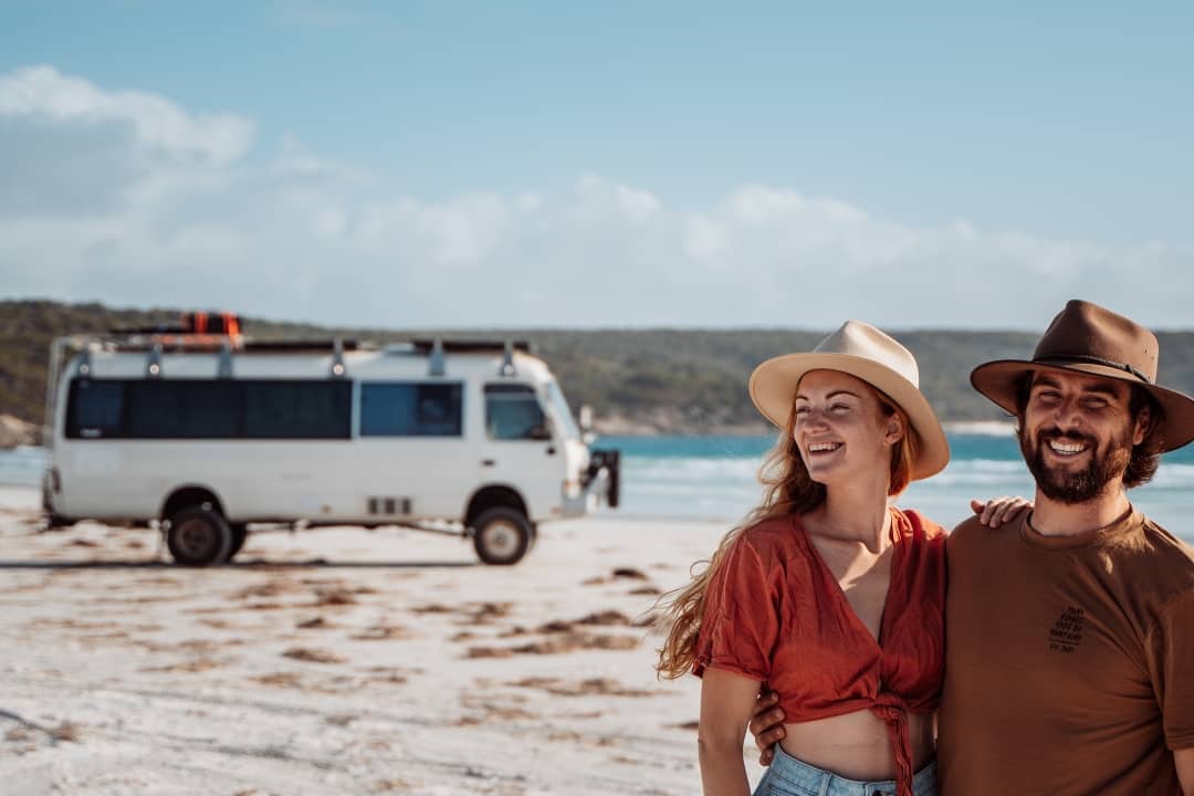 A smiling couple poses on a beach with a white van in the background, enjoying the beach in Bremer Bay Western Australia.