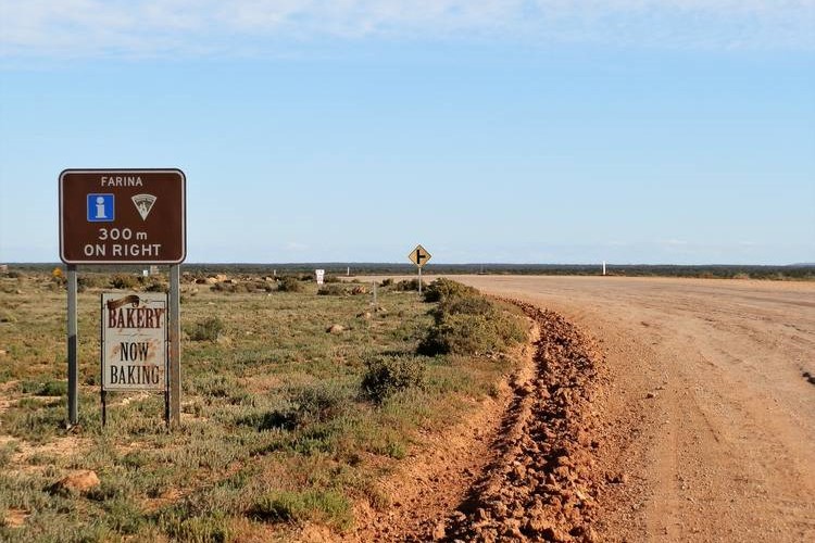 A sign by a dirt road in Farina, Outback South Australia, indicates that the farina outback bakery is "Now Baking." The sign also shows the direction to the town, 300 meters on the right.