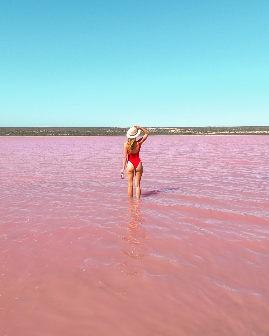 A woman in a red swimsuit stands in the pink waters of Hutt Lagoon. This shows things to do in Kalbarri, Western Australia.