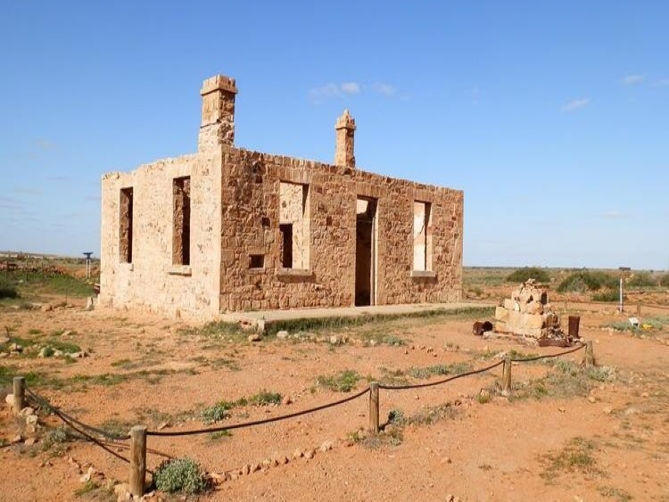 A photo of a ruined stone building in Farina, Outback South Australia, under a clear blue sky. The structure, surrounded by a simple rope barrier, stands on dry, sandy ground.