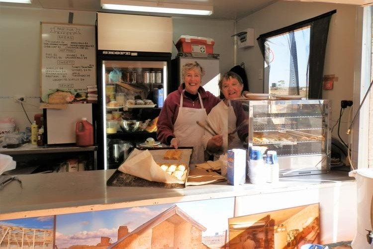 Two elderly smiling women serve fresh baked goods at a counter in the Farina Outback Bakery, South Australia. The bakery has a cozy interior with baked treats on display and a menu board in the background.