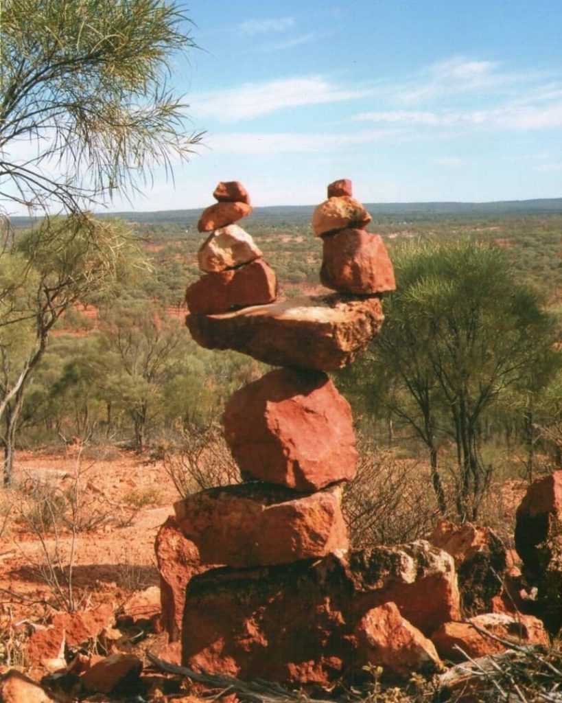 A scenic view from Yowah Lookout in Queensland, Australia, highlighting a balanced stack of reddish-brown rocks against a backdrop of dry, sparse vegetation and a distant horizon under a blue sky.