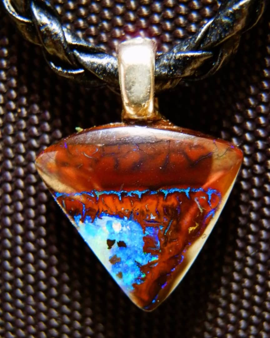 A close-up of a Yowah nut pendant, showcasing vibrant patterns and colors. The pendant features a mix of earthy brown tones with vivid blue opal inclusions, suspended from a braided black leather cord.