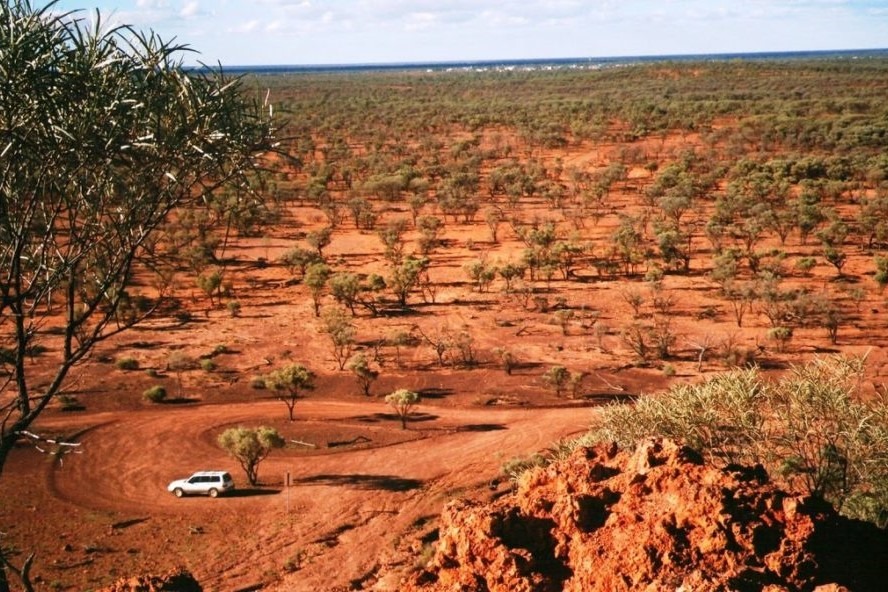 Aerial view of the arid outback of Quilpie, Queensland, with white SUV on red dirt road surrounded by sparse vegetation and open landscape.