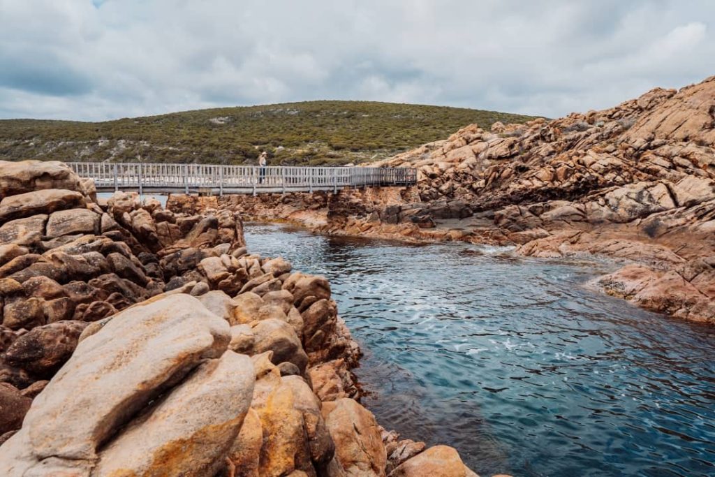 Canal Rocks in Margaret River, WA, featuring a wooden bridge over calm water surrounded by rugged orange-brown rocks and green hills in the background.