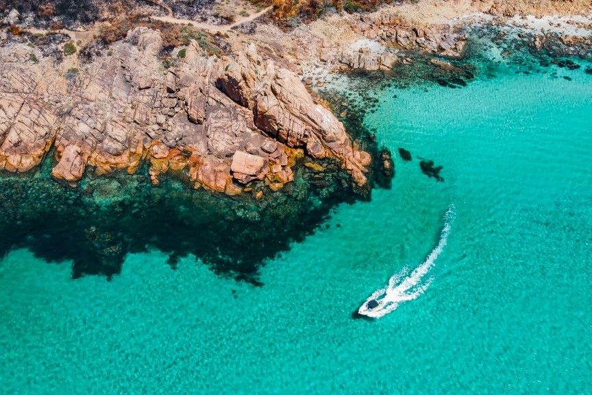 Aerial view of Castle Rock near Dunsborough, Western Australia, showcasing rugged reddish-brown cliffs meeting clear turquoise waters with a small boat cruising along the serene coastline. Perfect for those exploring things to do in Dunsborough.