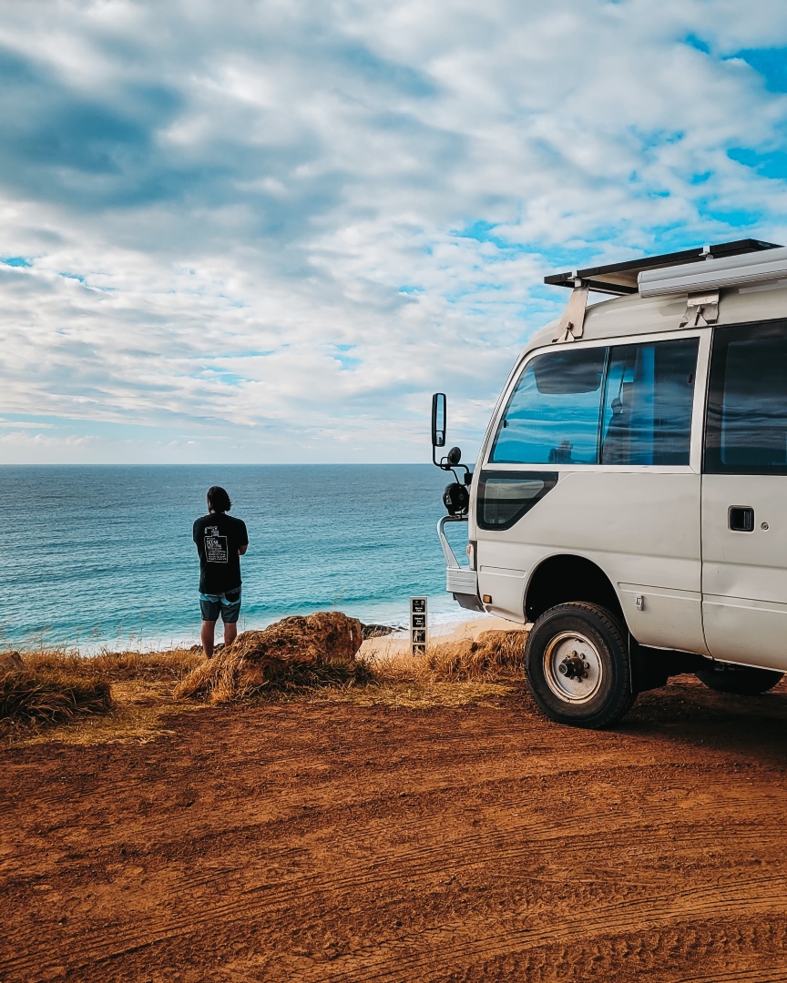 A person stands by the ocean, looking out over the water next to a parked camper van in the Injidup area near Yallingup, WA, with a cloudy sky overhead.