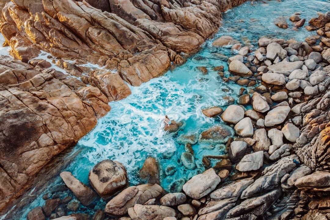 An aerial view of the Injidup Natural Spa, featuring large orange-brown rocks surrounding a small inlet of bright blue, foamy water. The scene is both rugged and serene, with waves crashing between the rocks.