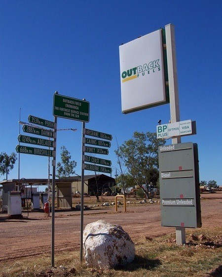 An outback fuel station with signs showing distances to cities like Perth and Darwin, set in a rugged, dry landscape under a clear sky.