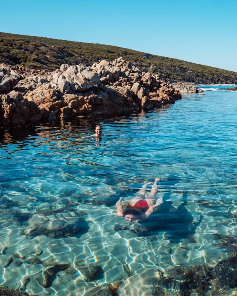 Two people swimming in the clear waters of The Aquarium Yallingup, surrounded by rocky shores and green hills.