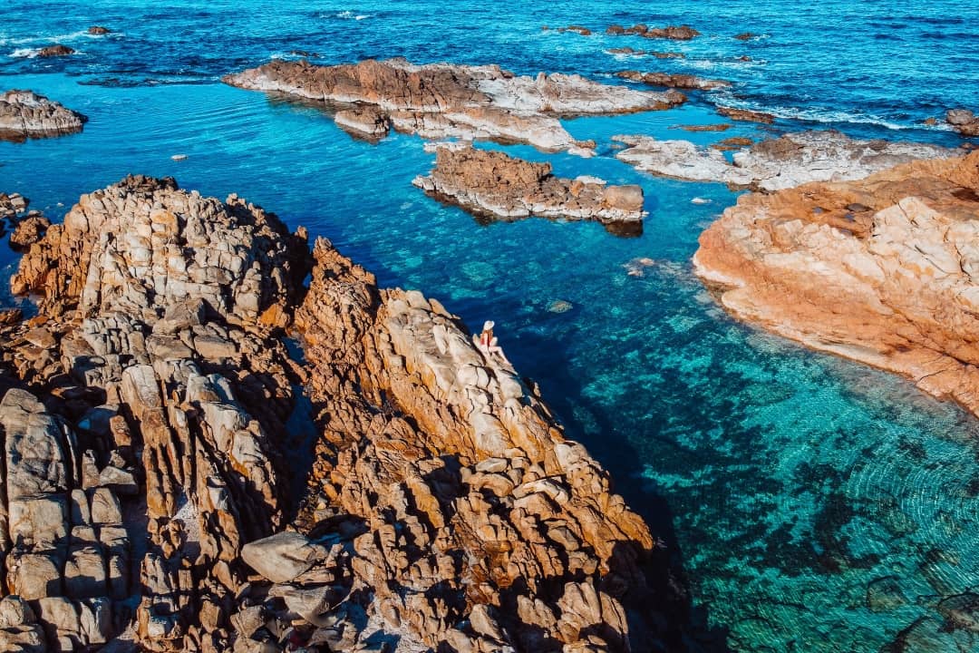 A rocky coastline at The Aquarium Yallingup with jagged orange-brown rocks jutting into clear turquoise water. A person sits on the rocks, enjoying the serene view.