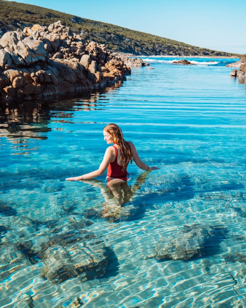 A woman in a red swimsuit wades into the clear turquoise waters of The Aquarium Yallingup, Western Australia, surrounded by rocky shores and hills.