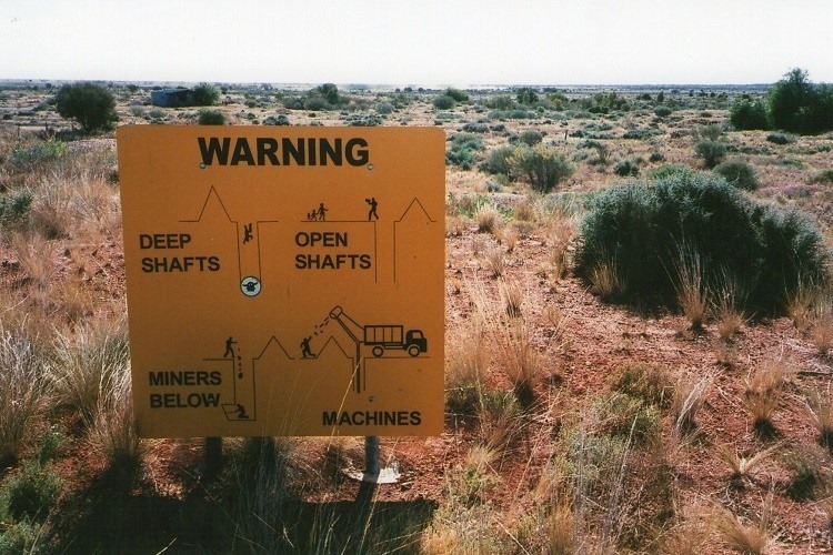 Yellow warning sign in a desert landscape near Quilpie, Queensland, cautioning about deep shafts, open shafts, miners below, and operating machines when mining for opals in Australia