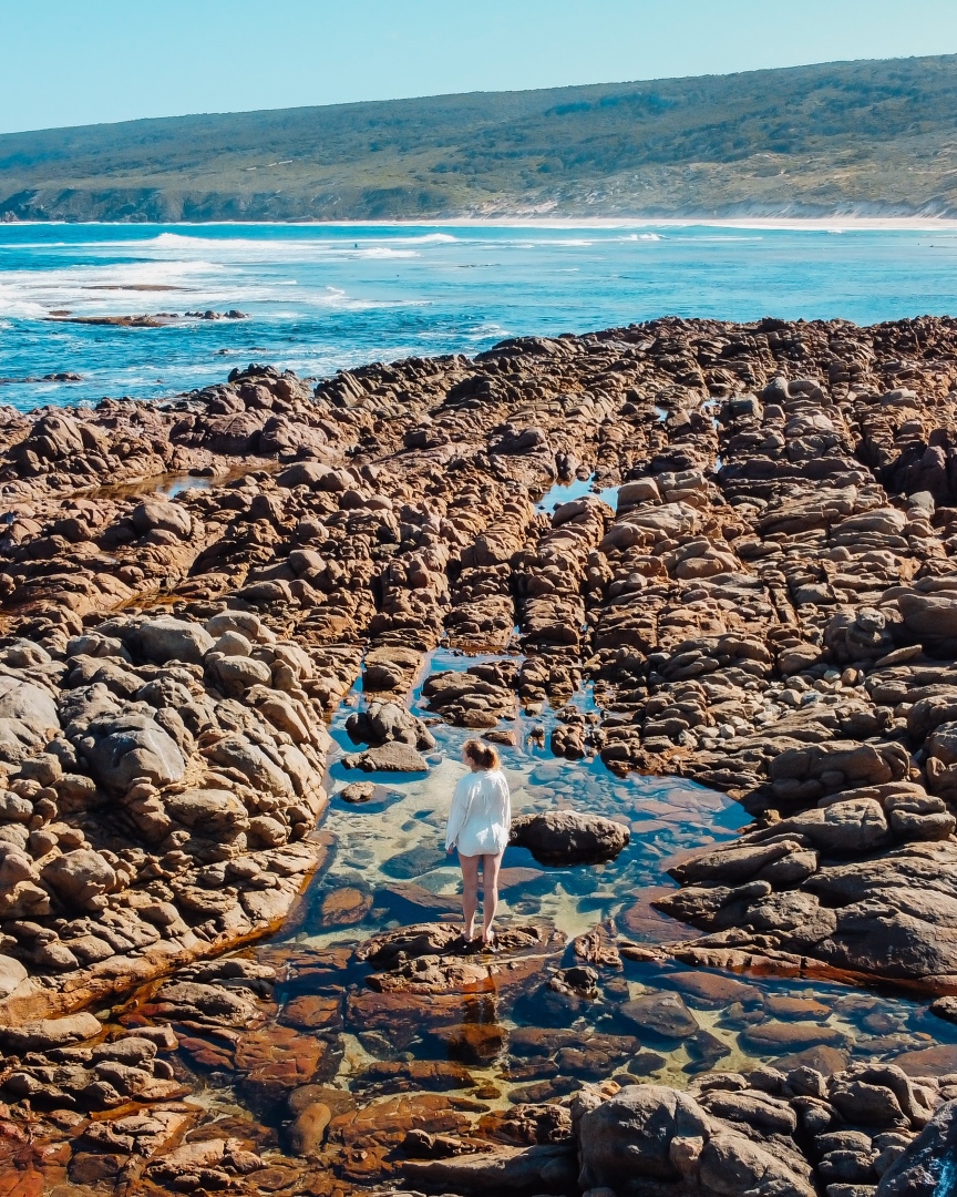 A person stands on rocky terrain near Yallingup, WA, surrounded by tidal pools with the ocean and hills visible in the background.