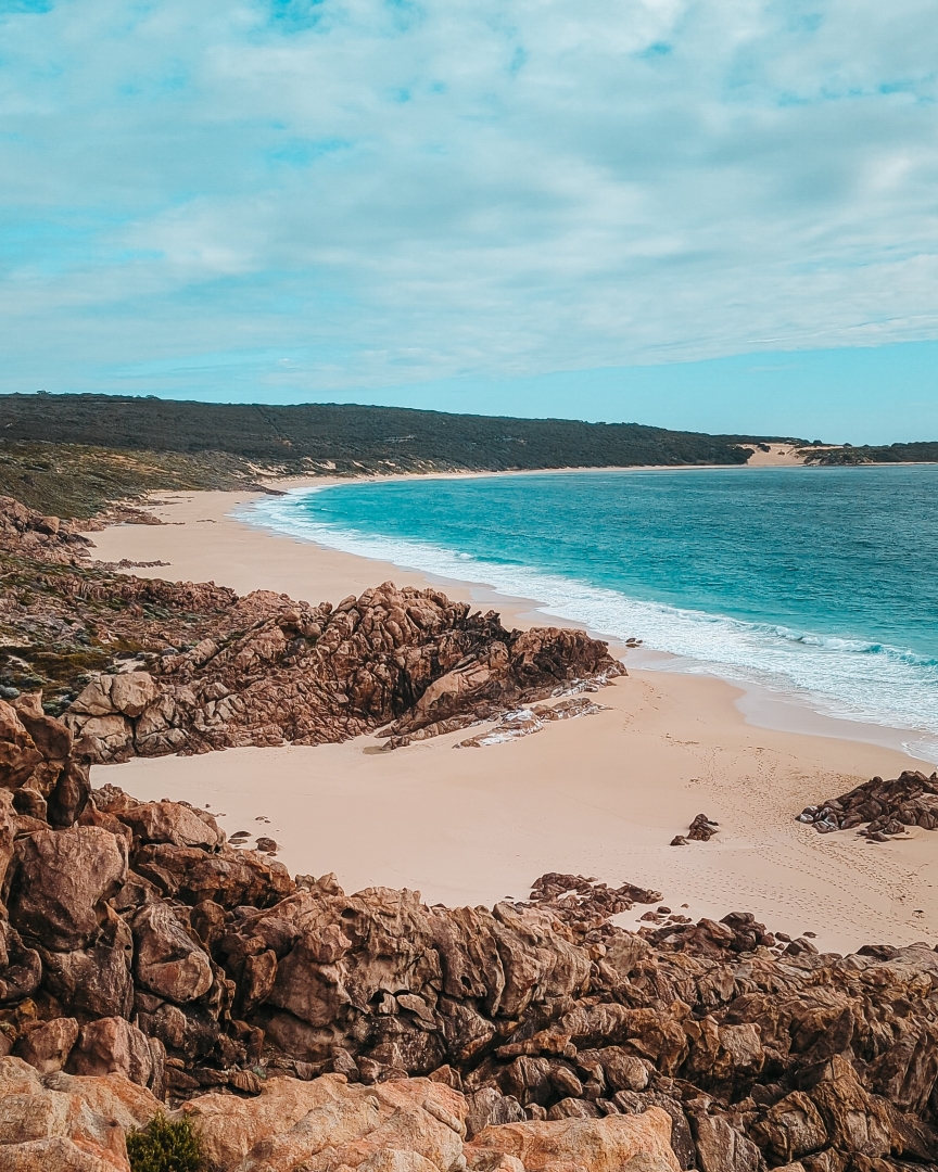 A sandy beach near Yallingup, WA, with rocky formations, turquoise waves, and hills in the background.