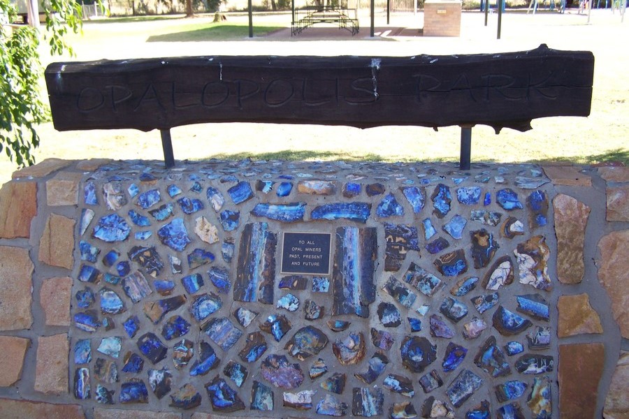 A memorial at Opalopolis Park dedicated to opal miners, featuring a plaque that honors miners past, present, and future, embedded within a stone wall adorned with colorful opal pieces.
