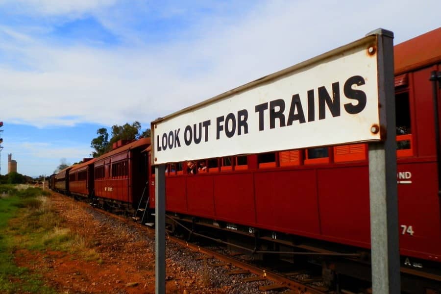 A sign reading "LOOK OUT FOR TRAINS" stands prominently in the foreground, with a vintage red train passing by on the tracks in Quorn, part of the Pichi Richi Railway, under a partly cloudy sky.