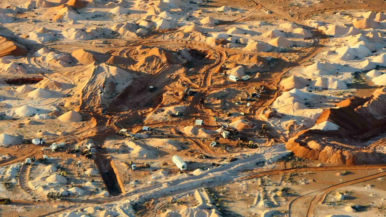 Aerial view of where to find opals in Australia. It shows a mining site in Coober Pedy, South Australia, with numerous excavation mounds and mining equipment spread across the arid, red desert landscape.