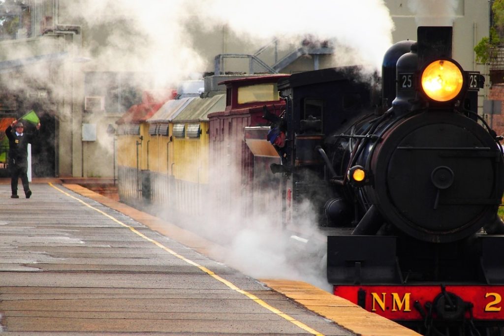 Pichi Richi steam train at a station, emitting clouds of steam, with a conductor signaling on the platform. Discover things to do in Port Augusta, including riding this historic steam train.