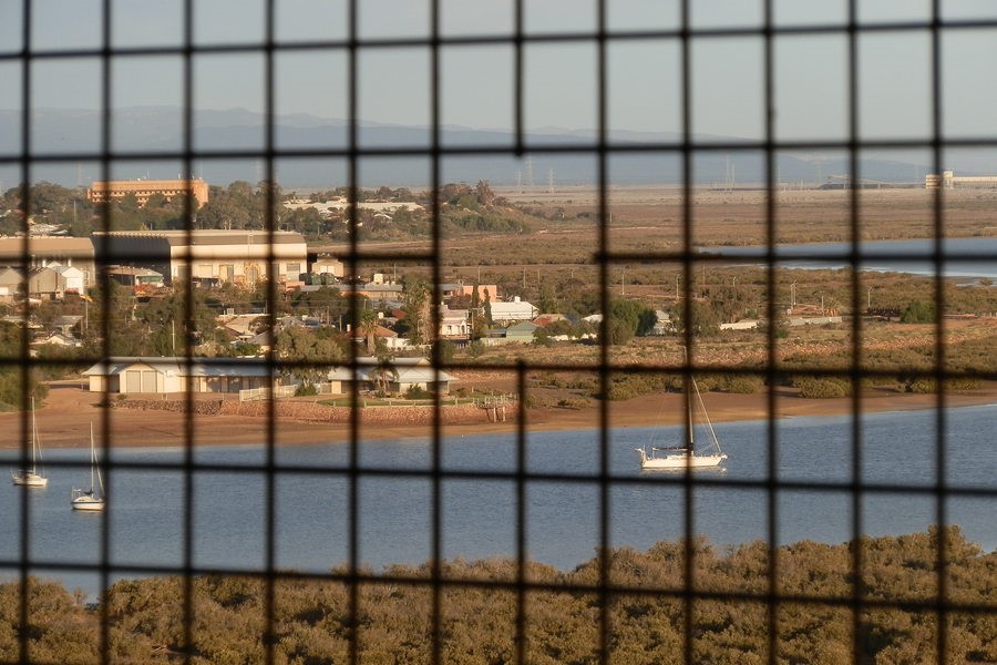 View of Spencer Gulf and Port Augusta from the water tower lookout, framed by a metal grid, with boats on the water and buildings in the background. Discover things to do in Port Augusta from this scenic vantage point.