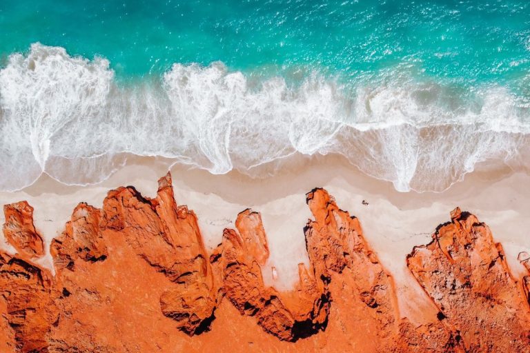 The secret Aussie hotspot of Cape Leveque, Broome, Dampier Peninsula: A striking aerial view of red sandstone cliffs meeting a white sandy beach with turquoise ocean waves crashing against the shore. The vibrant contrast of red rock and blue water creates a visually stunning natural scene.