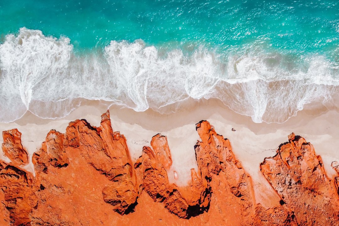 The secret Aussie hotspot of Cape Leveque, Broome, Dampier Peninsula: A striking aerial view of red sandstone cliffs meeting a white sandy beach with turquoise ocean waves crashing against the shore. The vibrant contrast of red rock and blue water creates a visually stunning natural scene.