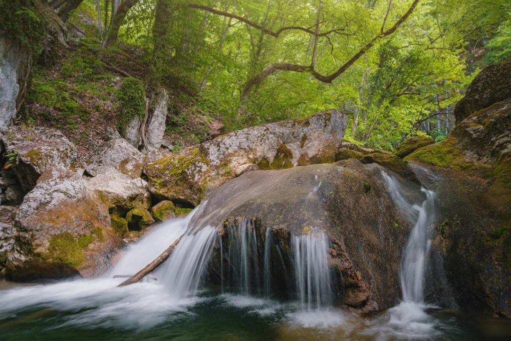 A small waterfall flows over large mossy rocks, surrounded by a lush green forest. The water cascades gently into a small pool, creating a peaceful nature scene.