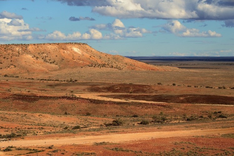 The Jump Ups, Sturt National Park, NSW. A vast outback landscape featuring flat-topped hills and red dirt plains under a partly cloudy sky. The contrasting colors of the desert terrain and the blue sky create a typical Australian outback view.