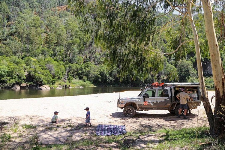 Jackson’s Crossing Beach, Snowy River, VIC. A tranquil riverside scene with a 4x4 vehicle parked on a sandy riverbank. Two children play near the water while another adult stands by the vehicle. The area is surrounded by lush greenery, making it a peaceful outdoor escape.