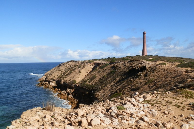 Troubridge Hill Lighthouse, Yorke Peninsula, SA. A lighthouse stands tall on a windswept cliff overlooking the ocean. The rocky coastline and clear blue sky highlight the isolated beauty of this coastal landmark.
