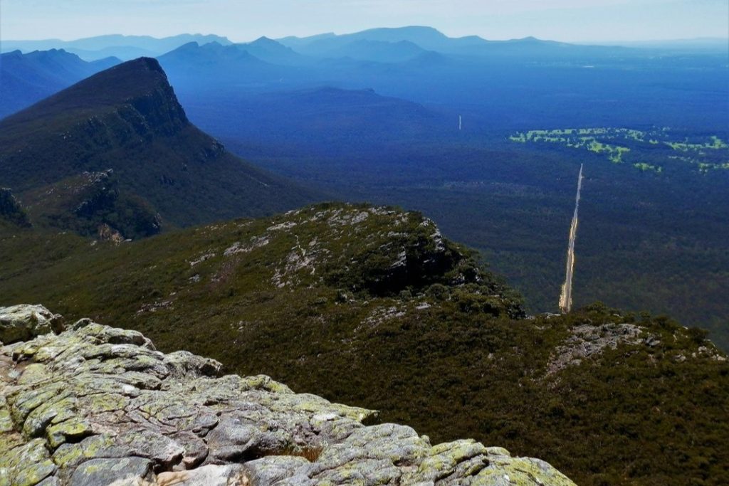Mt Abrupt, Grampians, VIC. A vast panoramic view from the top of Mt Abrupt, looking out over rolling mountains and dense forest below. The rugged rocky landscape contrasts with the distant hazy blue hills.