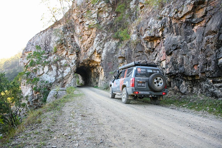 Old Glen Innes Road Tunnel, NSW. A dirt road leads to a historic tunnel carved into a rocky mountainside. The road is lined with cliffs, and the tunnel entrance appears rugged, showing the natural and man-made elements working together.