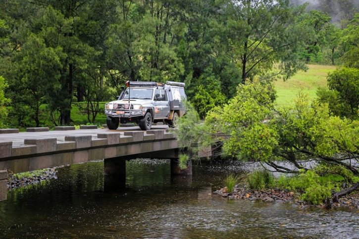 Old Glen Innes Road Bridge, NSW. A rugged off-road vehicle crosses a simple concrete bridge over a calm river, surrounded by greenery and trees. The bridge leads into a peaceful forest setting, suggesting adventure in a remote natural area.