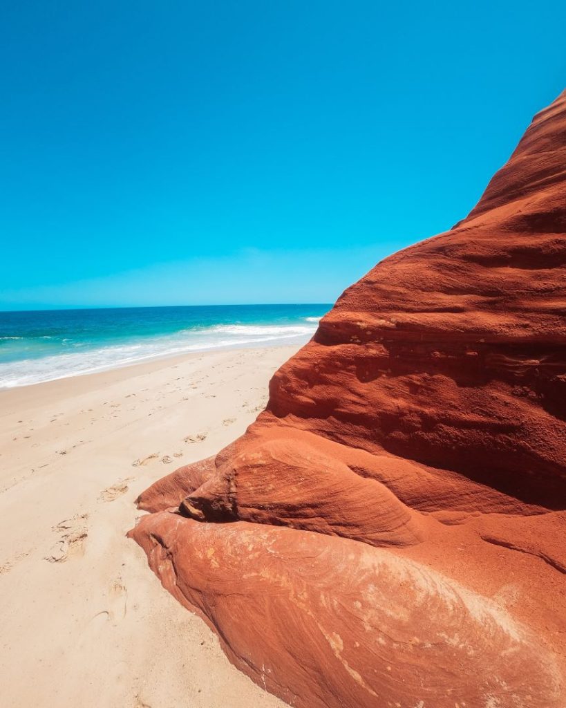 Cape Leveque, Broome, Western Australia. A vibrant red rock formation juts out onto a sandy beach with turquoise waves gently lapping the shore. Footprints trail along the sand, adding a human element to this serene coastal scene.
