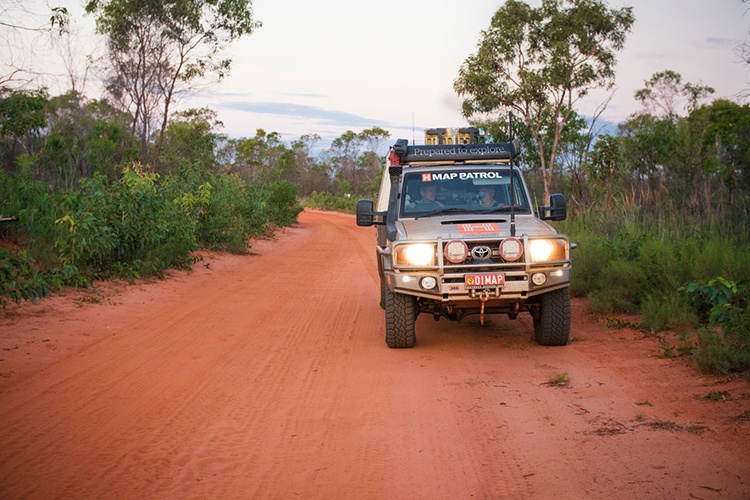 Gnylmarung Retreat, Cape Leveque. A 4x4 vehicle labeled "Map Patrol" drives along a red dirt road lined with green bushland. The scene reflects the remote and rugged nature of the Cape Leveque area, with tall trees and an open sky in the background. The dusty road contrasts with the lush vegetation, capturing the essence of an outback adventure.