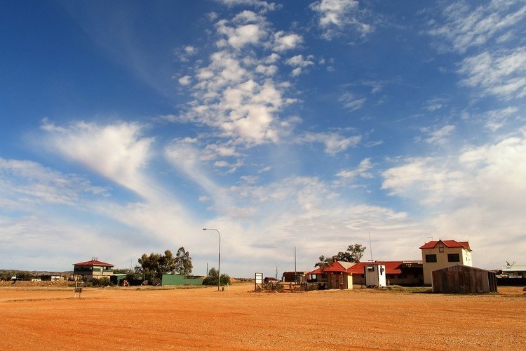 A few small buildings with red roofs on reddish dirt near Cameron Corner, under a vast blue sky with wispy clouds, as seen at the Alfred Hotel in Milparinka, NSW.