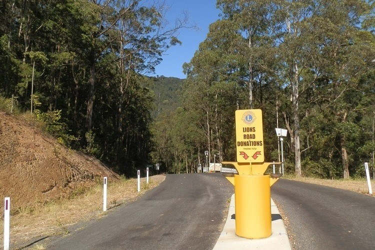 A yellow Lions Road donation station stands at the border crossing, surrounded by forest and hills.