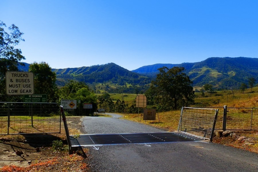 A scenic view of the Lions Road border crossing, featuring a cattle grid, road signs, and a backdrop of rolling green hills under a clear blue sky.