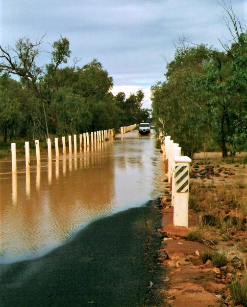 A flooded road crossing at the Bulloo River, with water covering the roadway and a vehicle approaching in the distance, reflecting typical outback conditions during heavy rains.