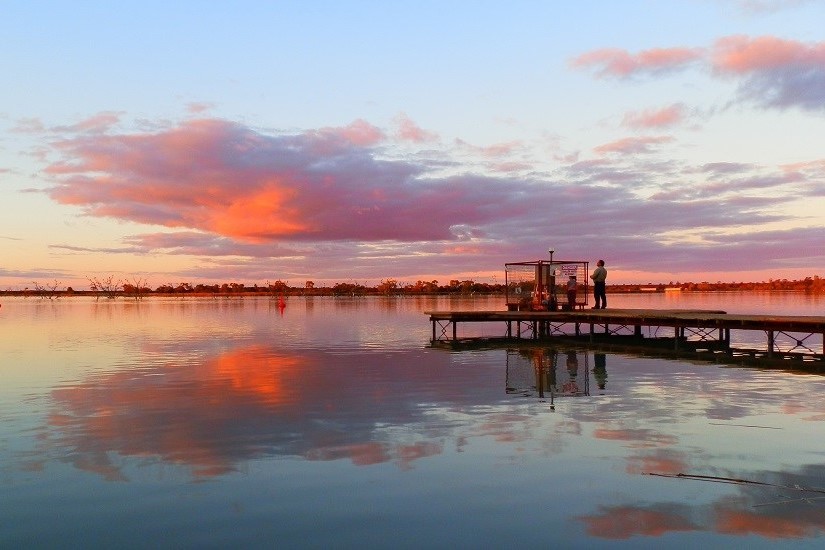 A person stands on the jetty at Lake Cullulleraine during sunset, with pink clouds reflecting on the calm water, showcasing serene activities among things to do at Lake Cullulleraine.